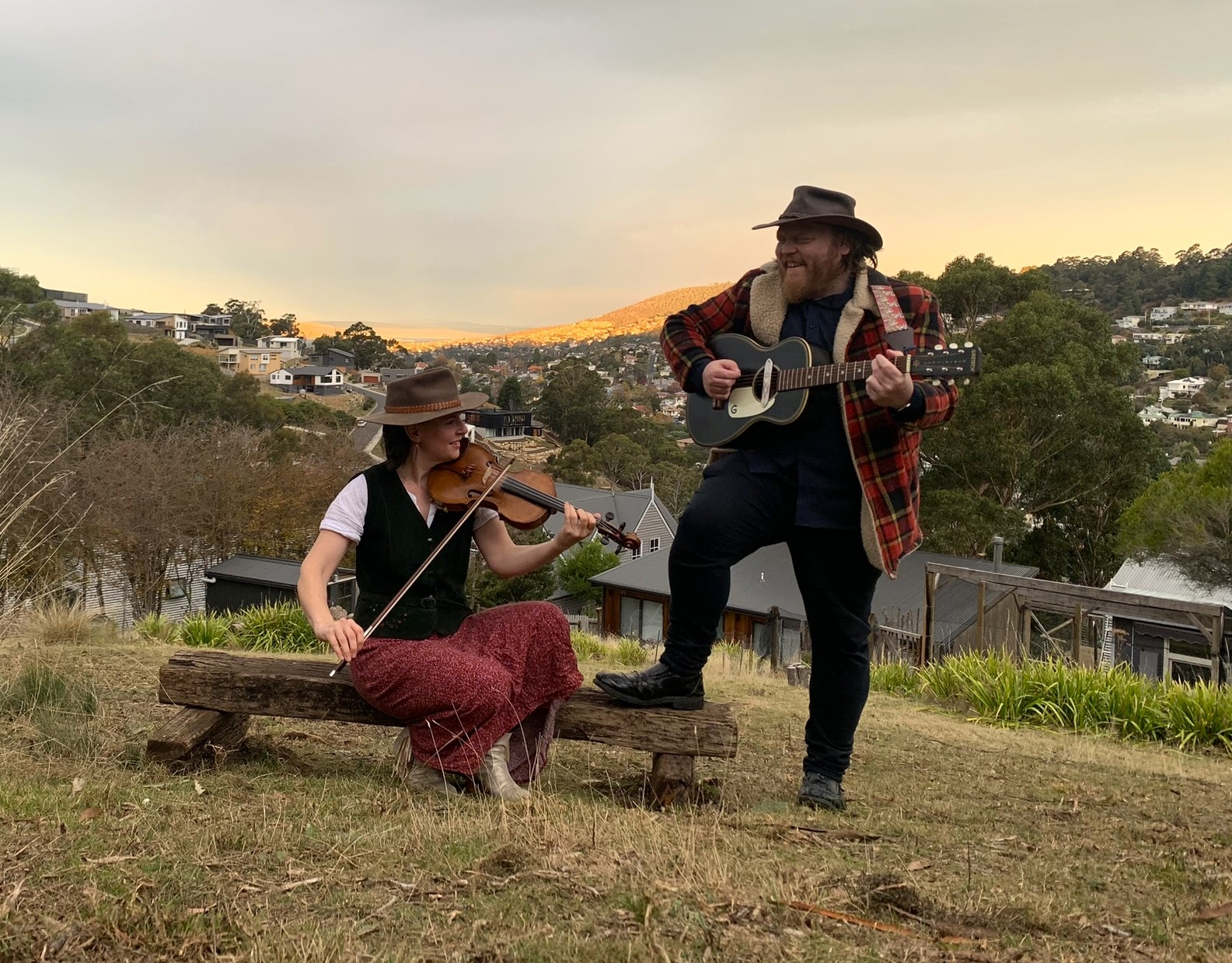 Outdoor photograph of Susannah sitting playing violin and Dave standing playing guitar